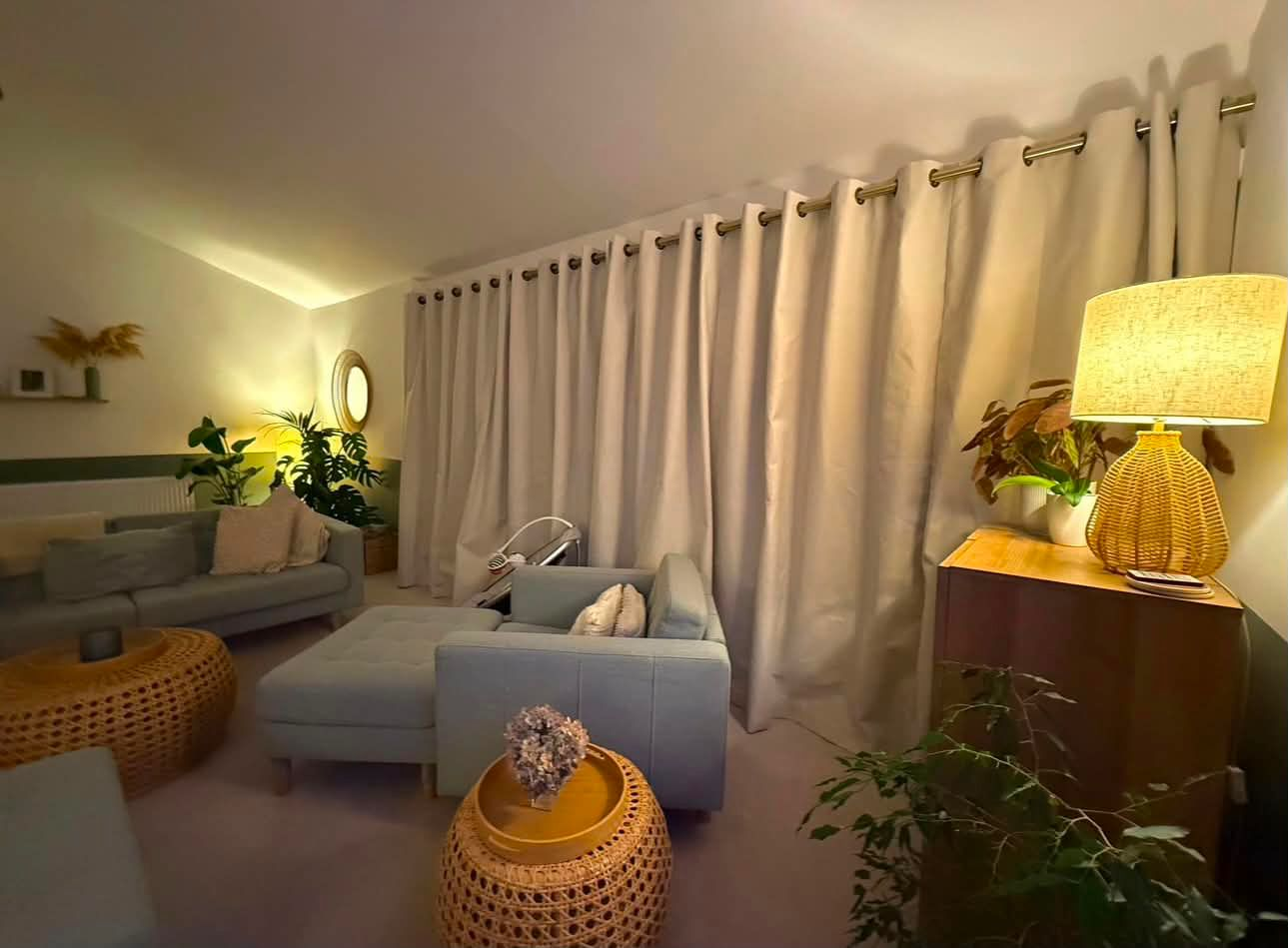 Cream eyelet curtains running across a wide lounge wall with a sofa and lamp