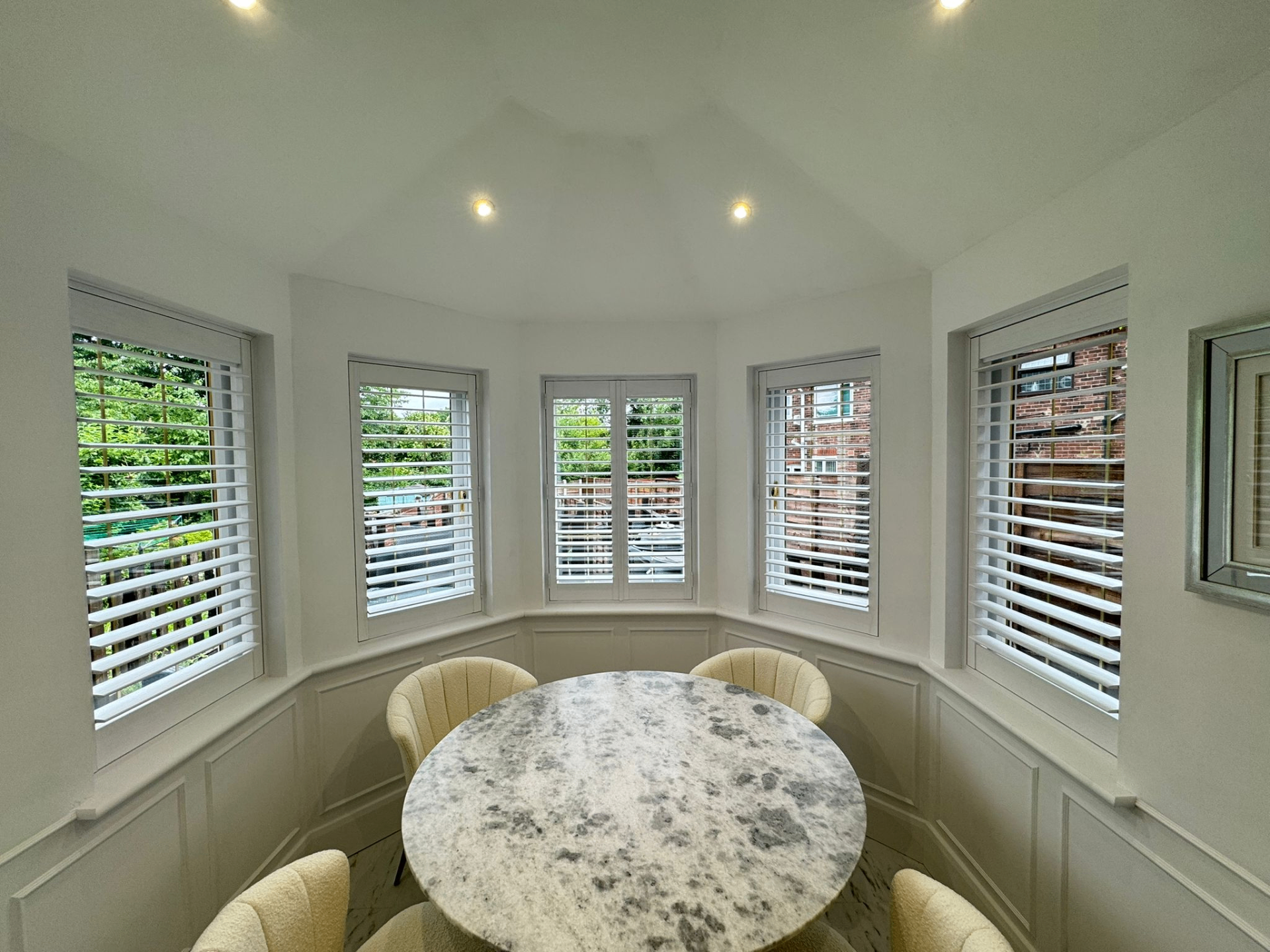 Full height white shutters fitted across a bright bay dining area with round marble table