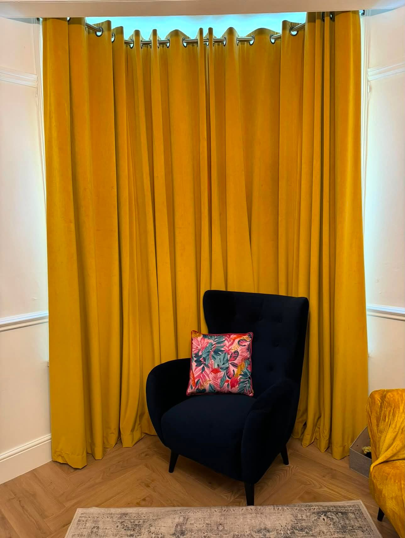 Mustard yellow velvet eyelet curtains across a bay window with a navy armchair