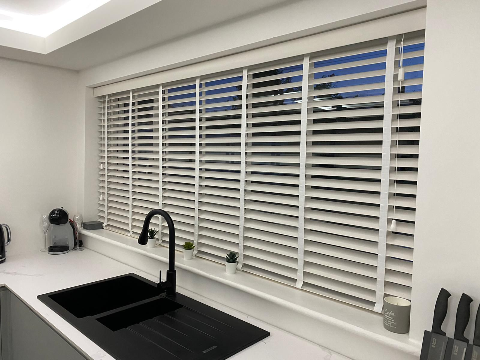 Wide white wooden Venetian blinds with white ladder tapes in a modern kitchen