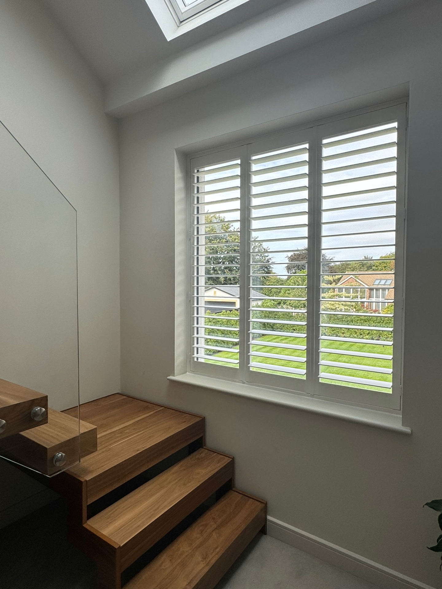 Full height white shutters fitted to a tall landing window beside timber stairs and rooflight
