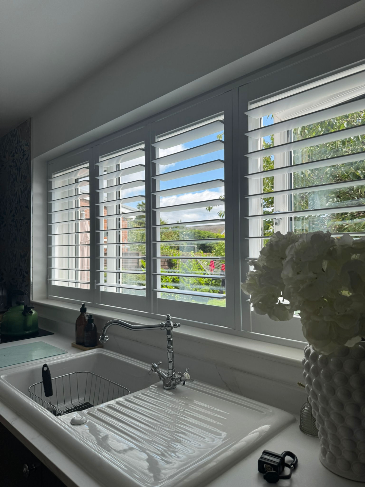 White PVC shutters fitted above a kitchen sink with garden views beyond